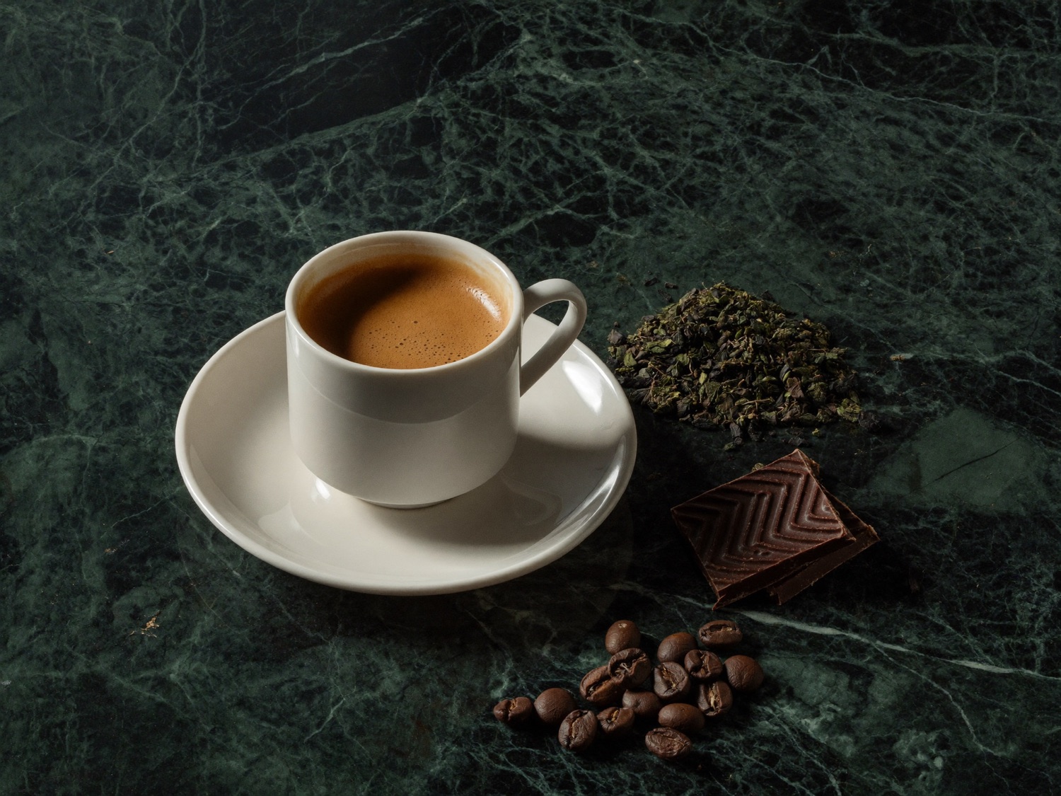 Tazza di espresso, foglie di te verde e quadratini di cioccolato fondente su un piano di marmo verde